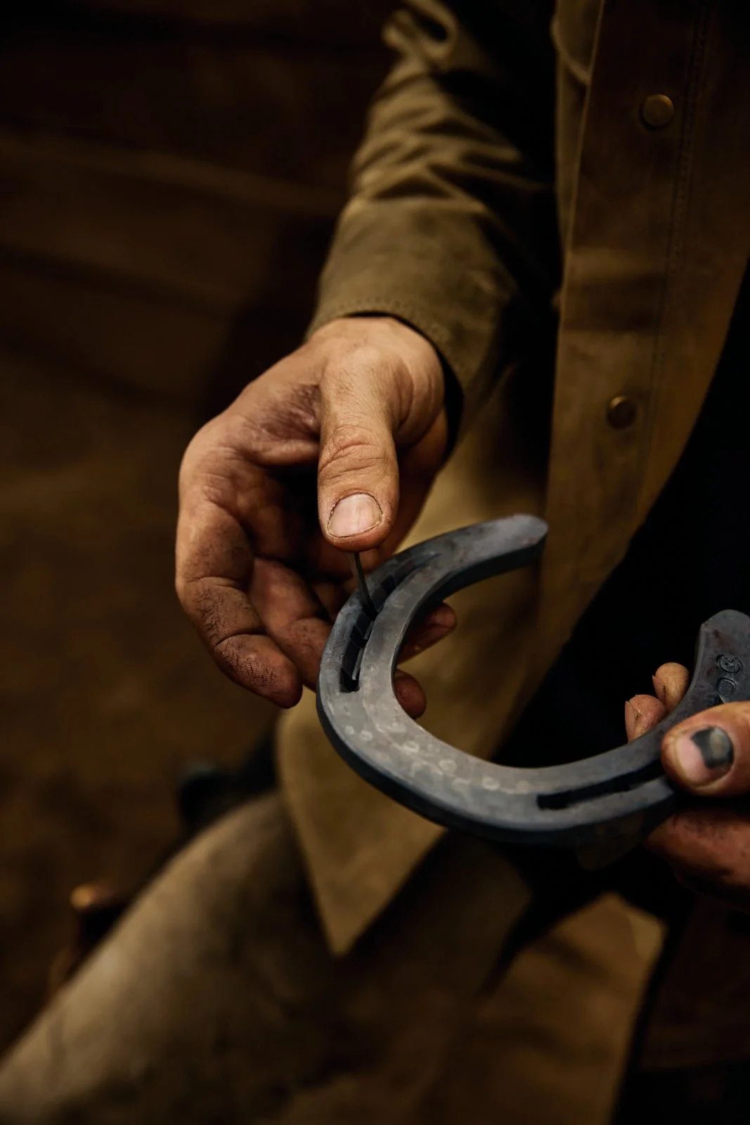 Close-up of a person holding a horseshoe, with one hand gripping the horseshoe and the other hand interacting with a tool, possibly for blacksmithing, with dirt and soot on their hands.