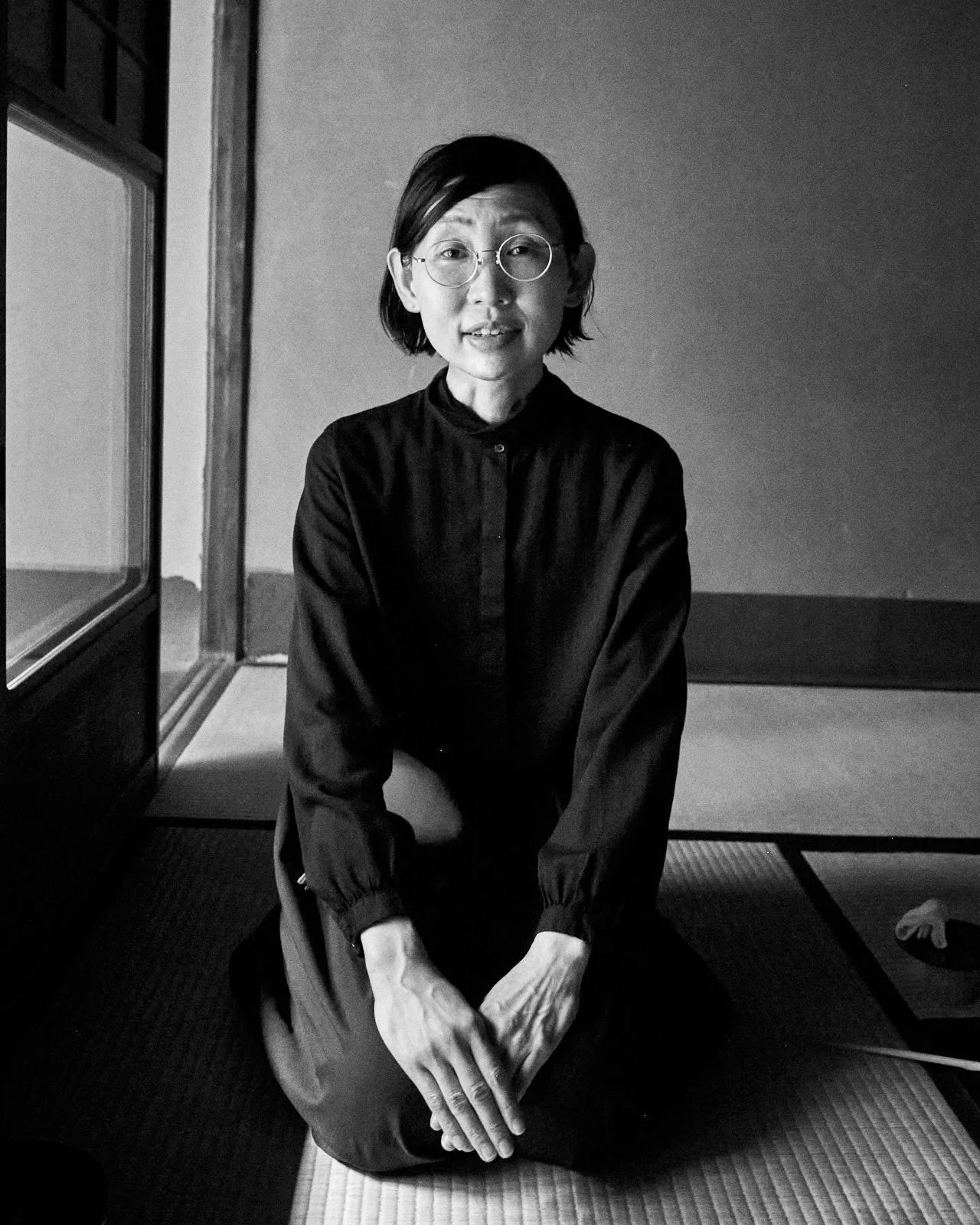 A woman with glasses and short dark hair sitting on the floor of a traditional Japanese room with tatami mats, looking at the camera.