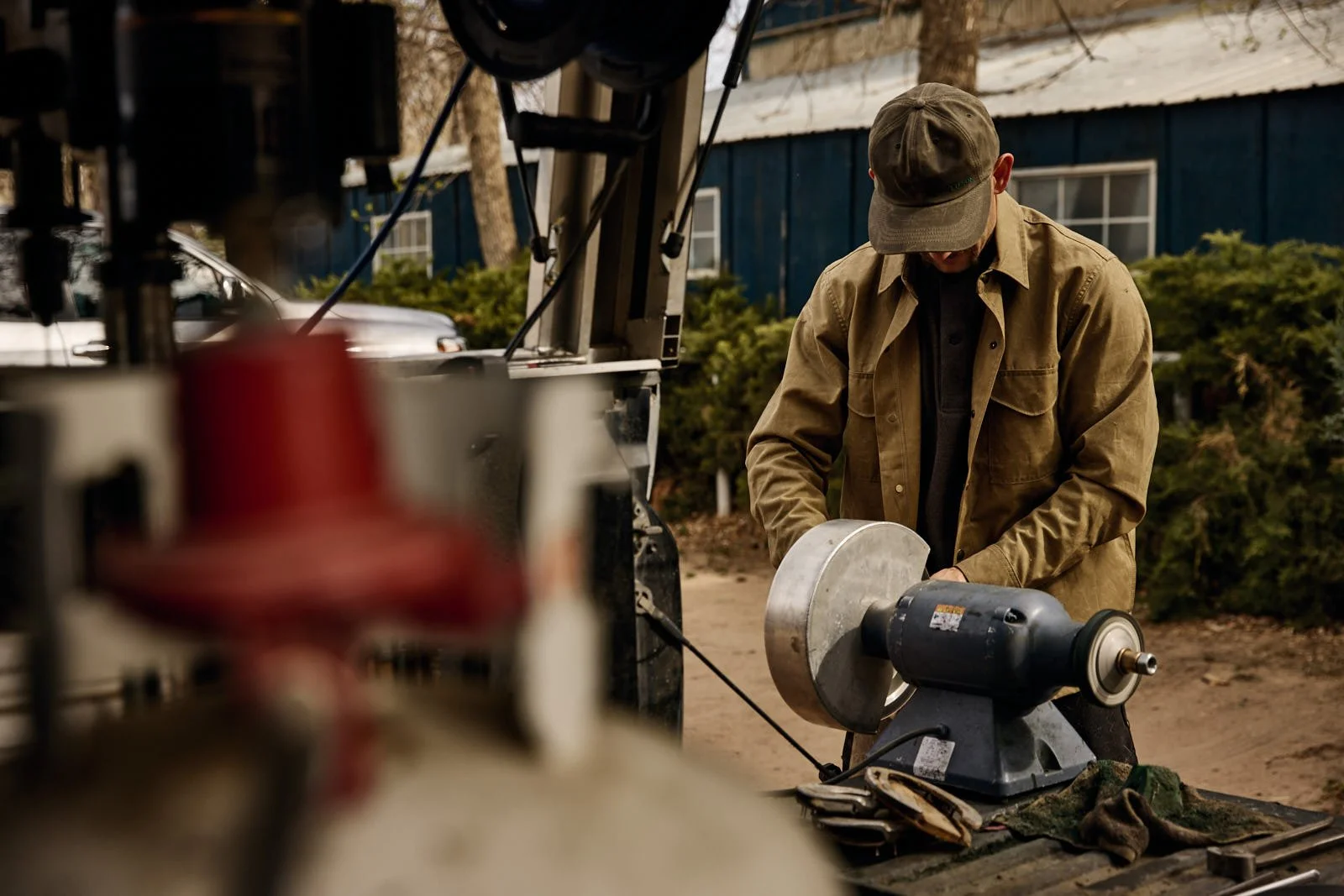 A man operating a bench grinder outdoors, wearing a brown jacket and cap, with a blurred machine in the foreground and a blue building in the background.