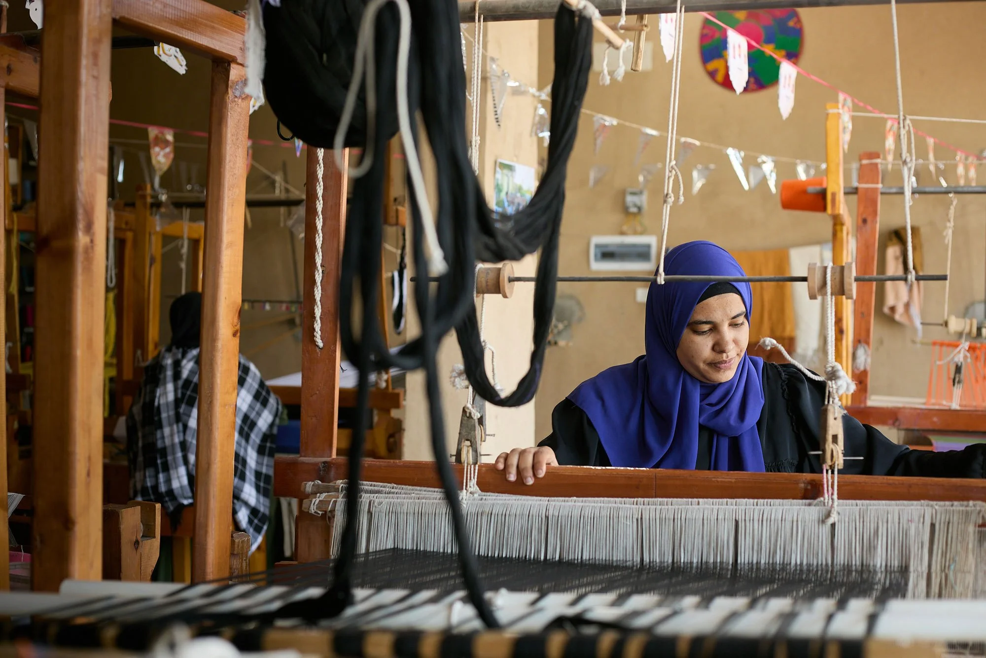 Two women, one wearing a blue hijab, are working on a traditional weaving loom inside a room decorated with colorful artwork and fabric. The room has wooden structures and hanging ropes for weaving.