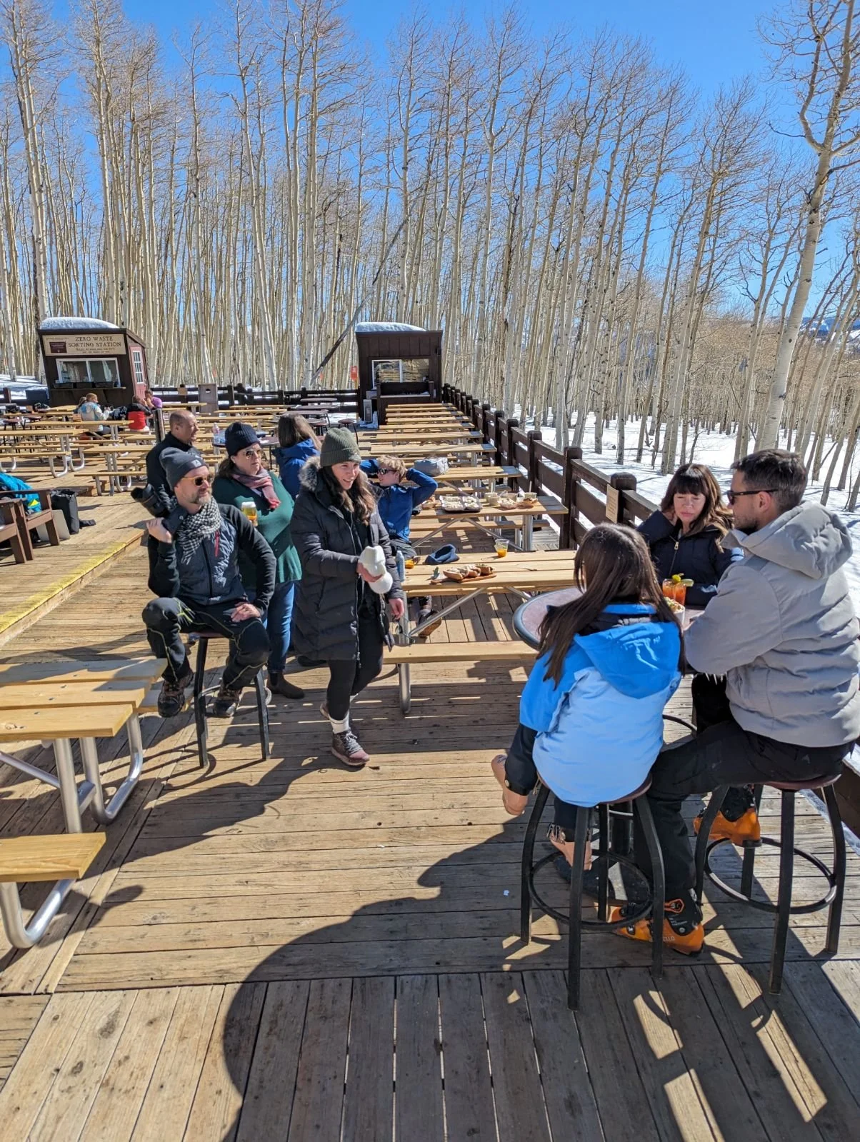 People gathered on an outdoor wooden deck with picnic tables, some standing and others sitting, wearing winter jackets and hats, with snow on the ground and leafless trees in the background at a Vail Resorts lodge in Breckenridge Colorado.