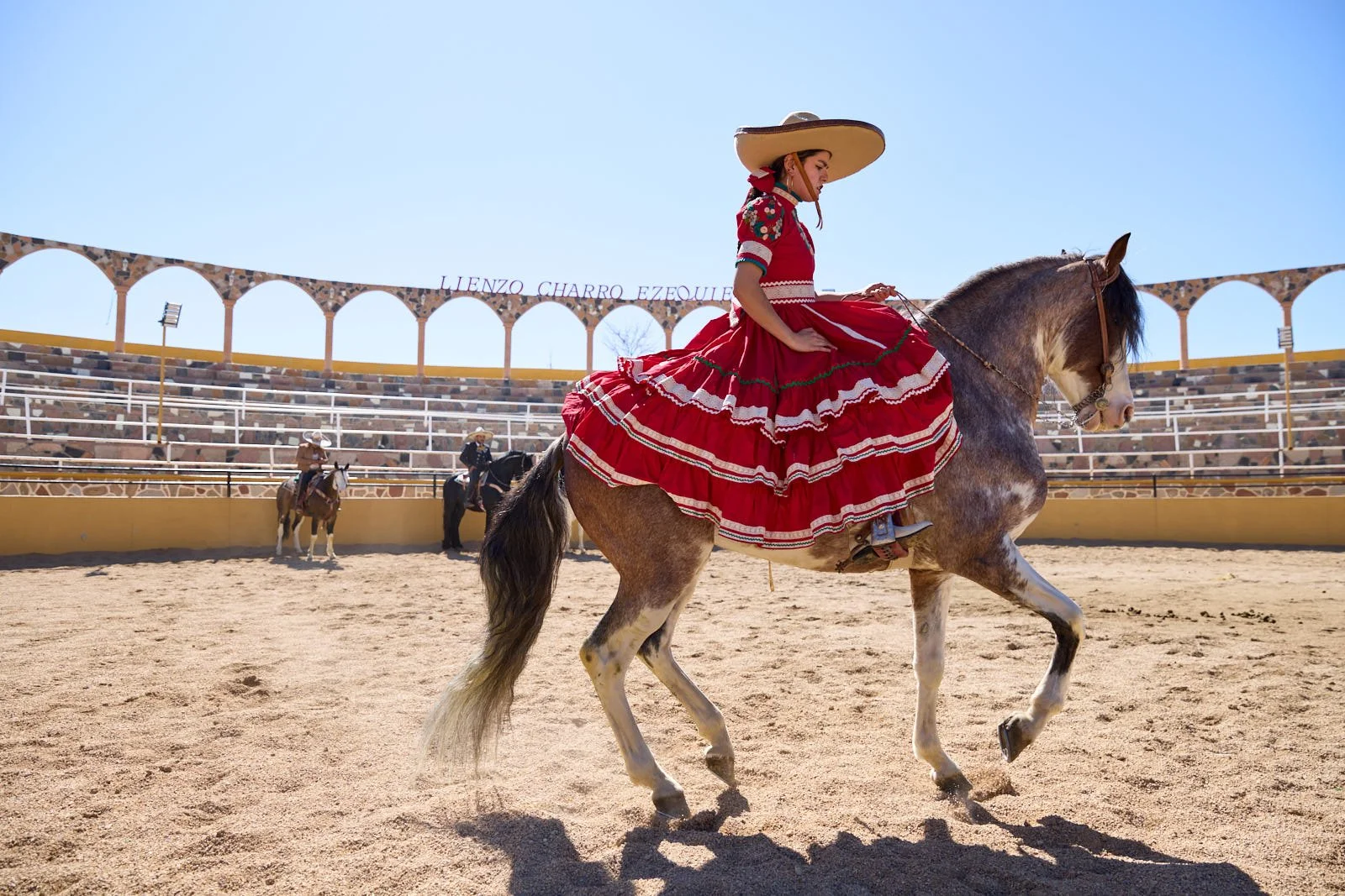 A woman in traditional Mexican attire riding a horse in an arena with tiered seating and a clear blue sky.