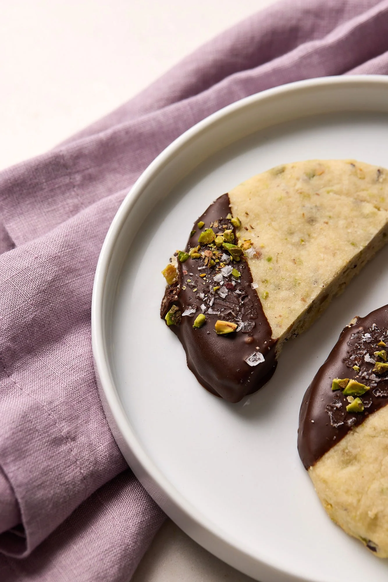Two chocolate dipped shortbread cookies on a white plate and purple napkin 