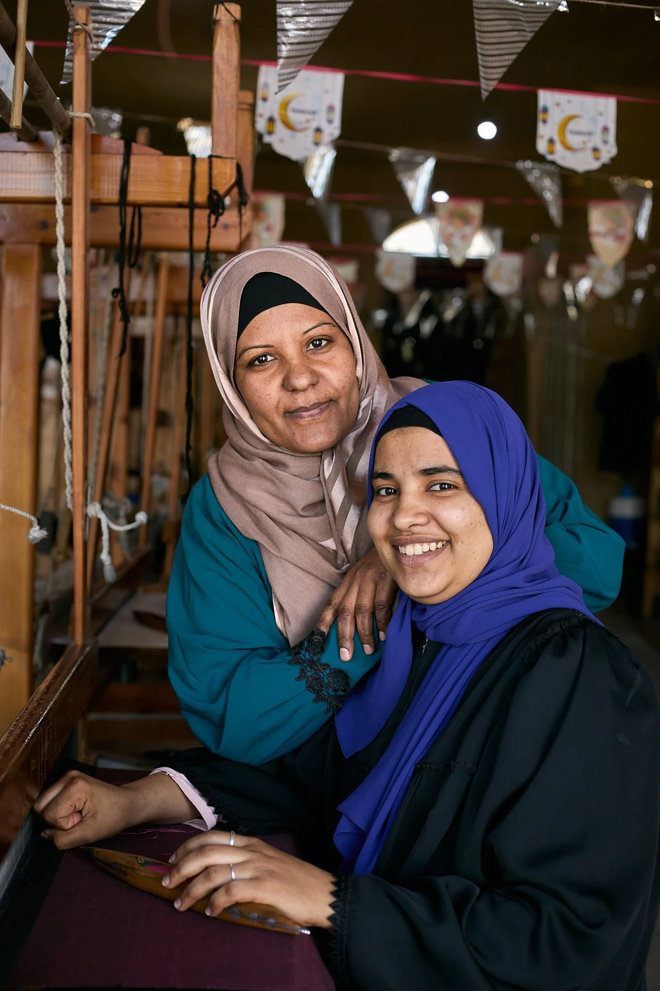 Two women in hijabs smiling and posing for a photo in front of a loom.
