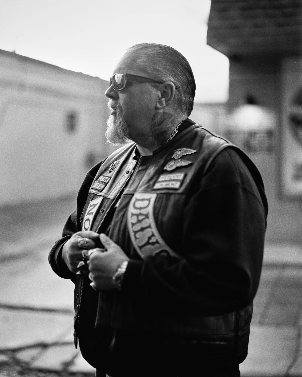 A black and white photograph of a man with a beard and glasses, wearing a leather vest with patches standing outdoors with blurred buildings in the background.