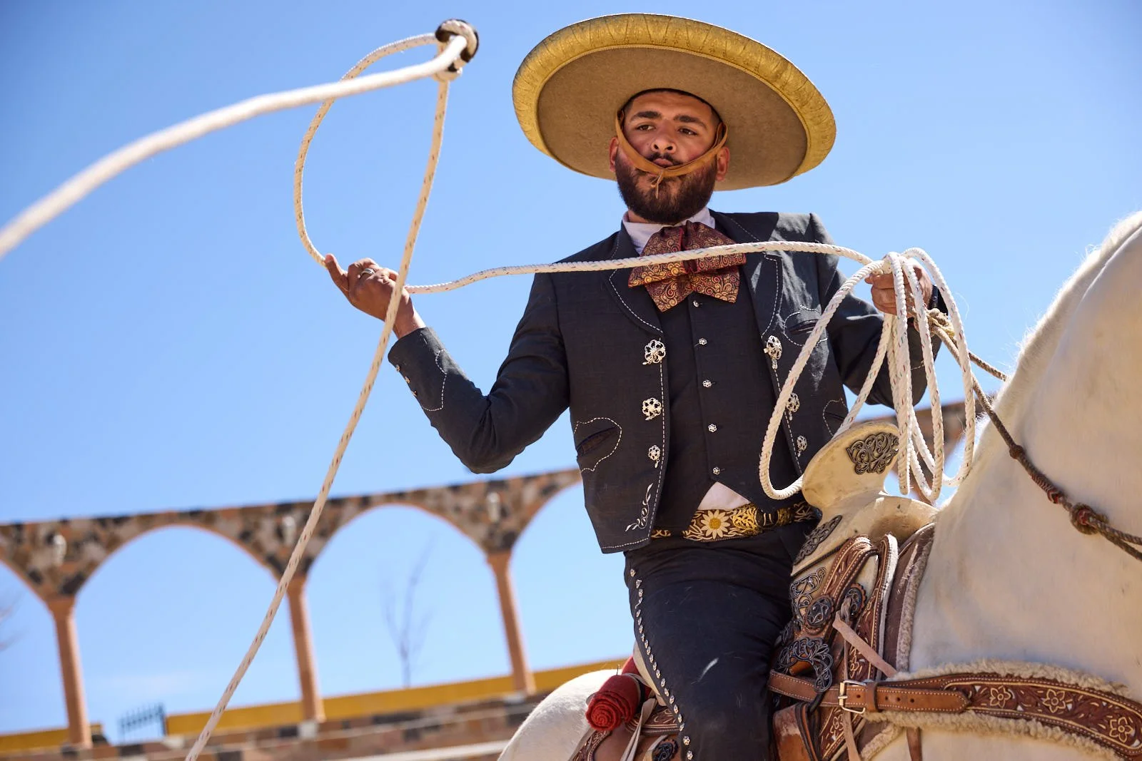 Man riding a white horse, wearing traditional Mexican attire including a sombrero, a black embroidered blazer, and a red neckerchief, holding a lasso against a clear blue sky.