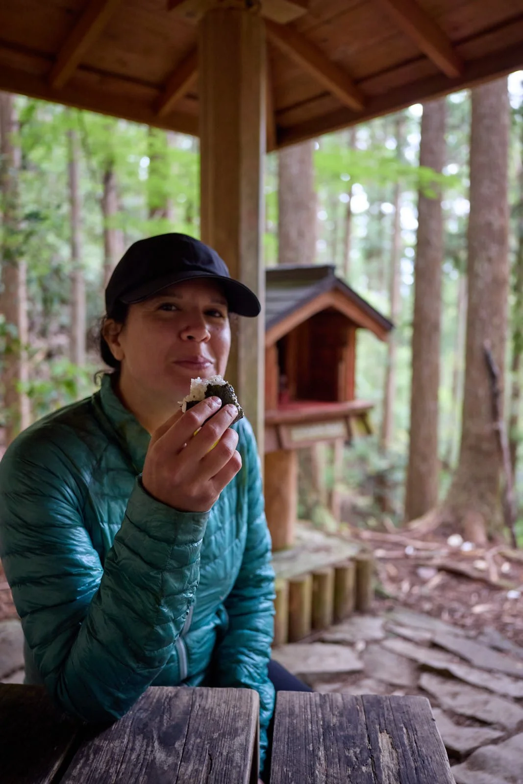 A woman wearing a black cap and teal puffy jacket is holding and eating a rice ball with seaweed in a forest setting under a wooden shelter.