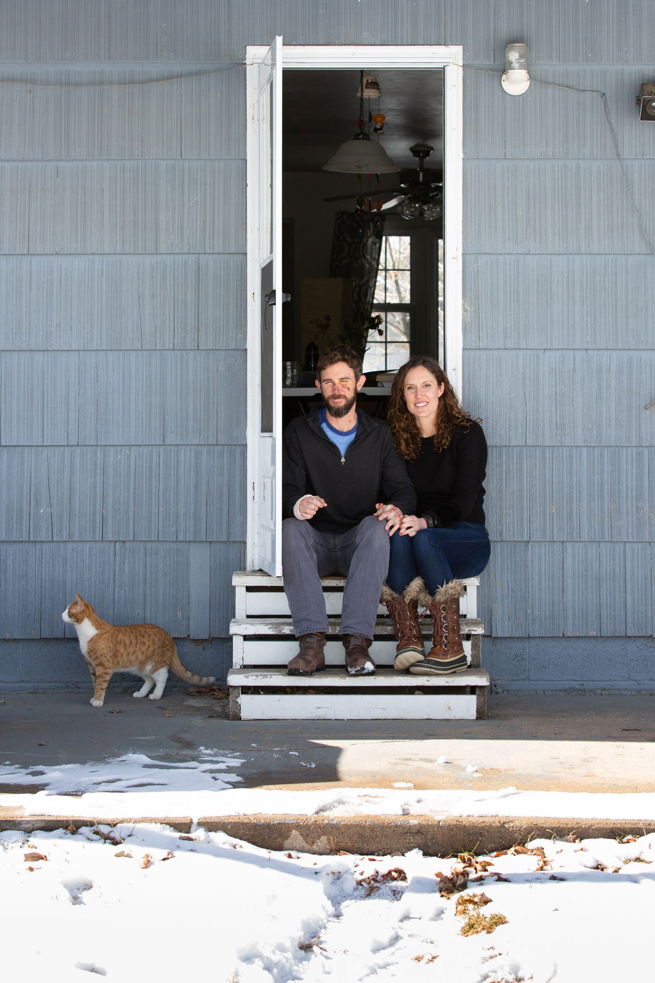 A couple sitting on front porch steps of a house, with a ginger cat nearby in the snow, in a winter setting.