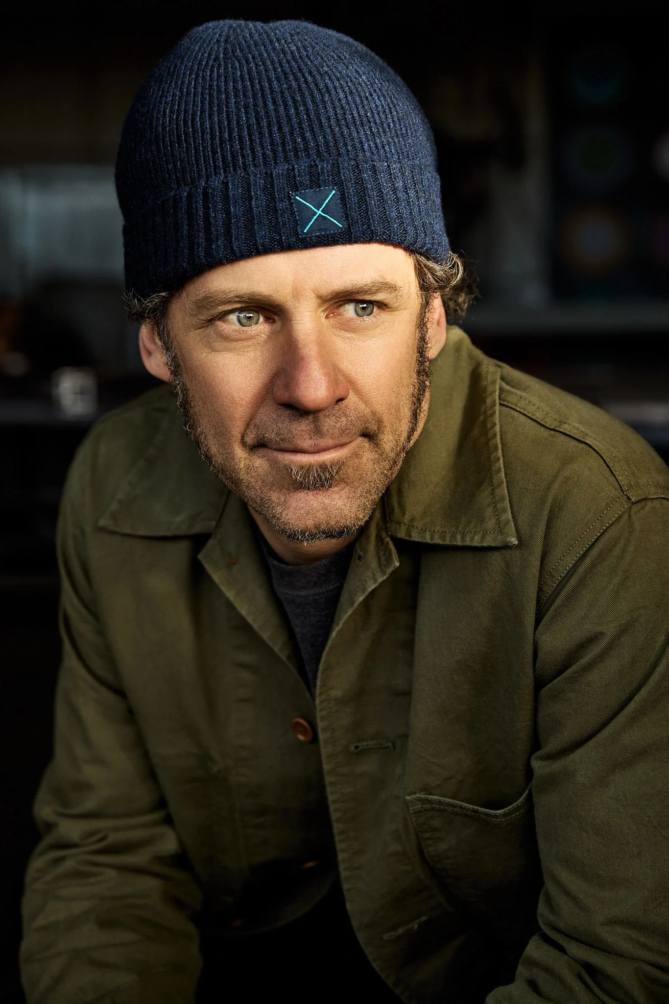 A man with grayish hair and a beard wearing a blue knit cap and an olive green jacket, looking to the side.