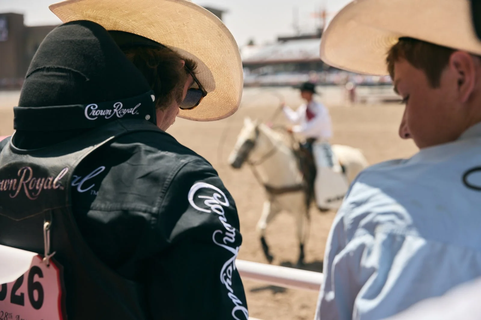 Two individuals wearing cowboy hats and Western clothing are seen from the back, with a person riding a horse in the background at a rodeo