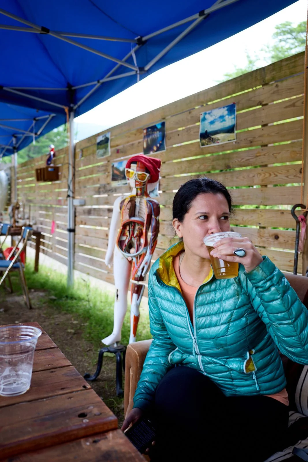 A woman in a teal jacket sitting outside at a wooden table, drinking beer from a clear plastic cup, with a skeletal anatomical model wearing sunglasses and a Santa hat in the background. There is a wooden fence with photos pinned on it and a blue can