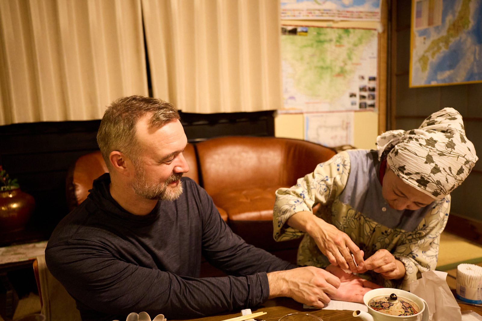 A man smiling while having his hand examined by a woman dressed in traditional clothing, sitting at a table with tea and other items, in a cozy room with maps and curtains in the background.