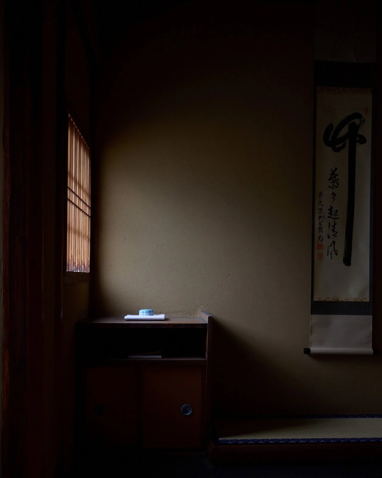 A dimly lit traditional Japanese interior with a paper window on the left, a small shelf with a rolled white cloth or paper and a small blue item on top, and hanging wall scroll with Japanese calligraphy on the right.