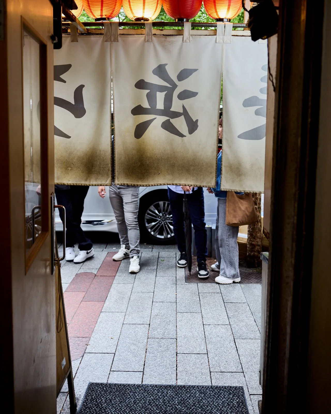 View through a restaurant doorway showing people standing outside, with a curtain featuring a Japanese character hanging overhead and red paper lanterns above.