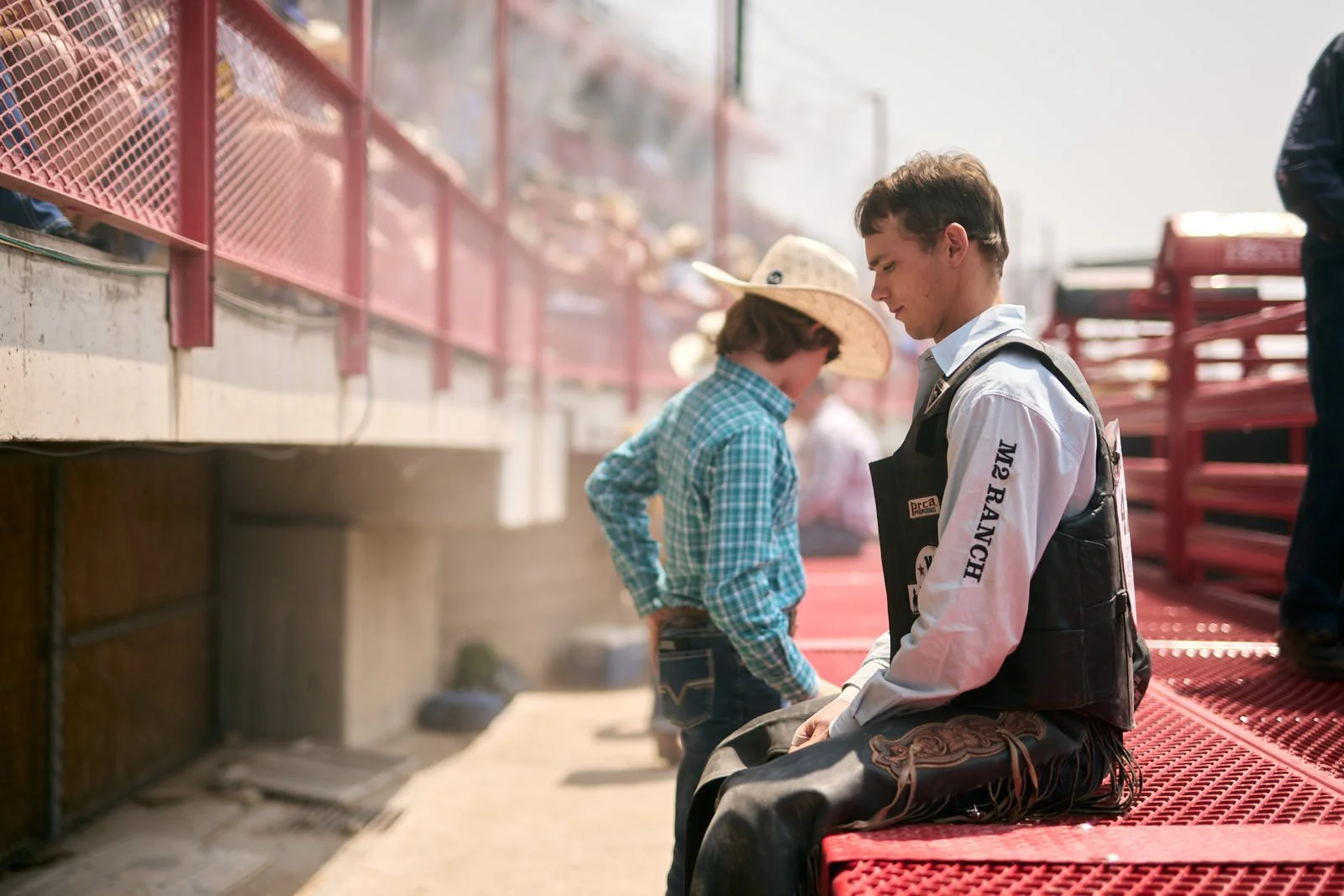 A young man dressed as a cowboy, sitting on a red metal railing at a rodeo.