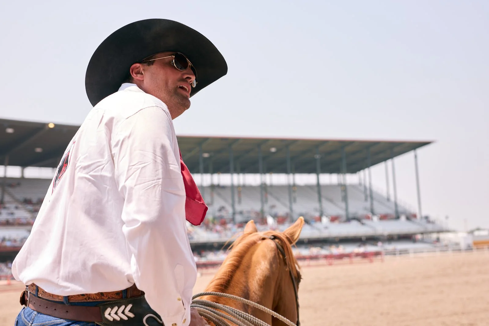 A man wearing a large black hat, sunglasses, a white shirt, and a red bandana riding a brown horse at a racetrack with a grandstand in the background.
