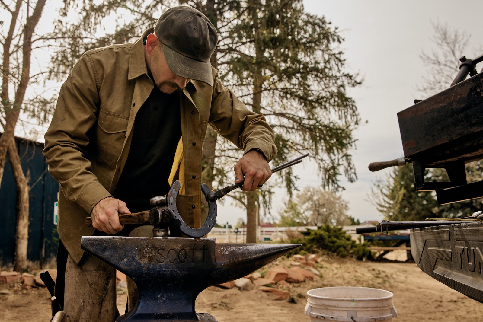 A man working outdoors with a hammer and horseshoe on an anvil, surrounded by trees and outdoor tools.