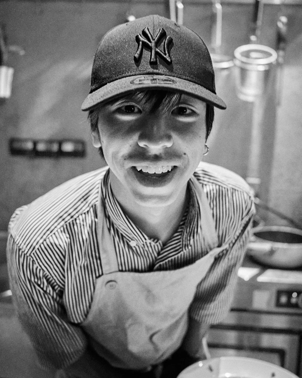 A smiling young person wearing a baseball cap and striped shirt, standing in a kitchen.