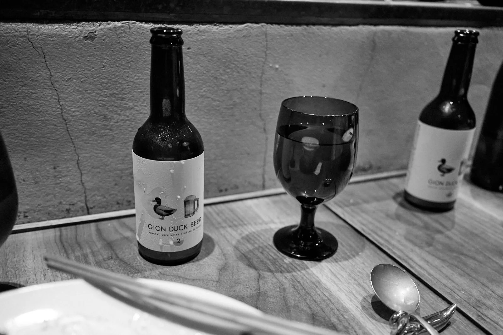 Bottles of Gion Duck Beer, a glass of dark beer, a spoon, and a bowl on a wooden table against a textured wall.
