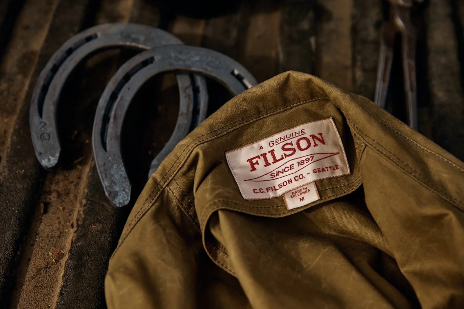 A brown Filson jacket with a red and white label resting on a rusty work surface, with two horseshoes and a pair of pliers nearby.