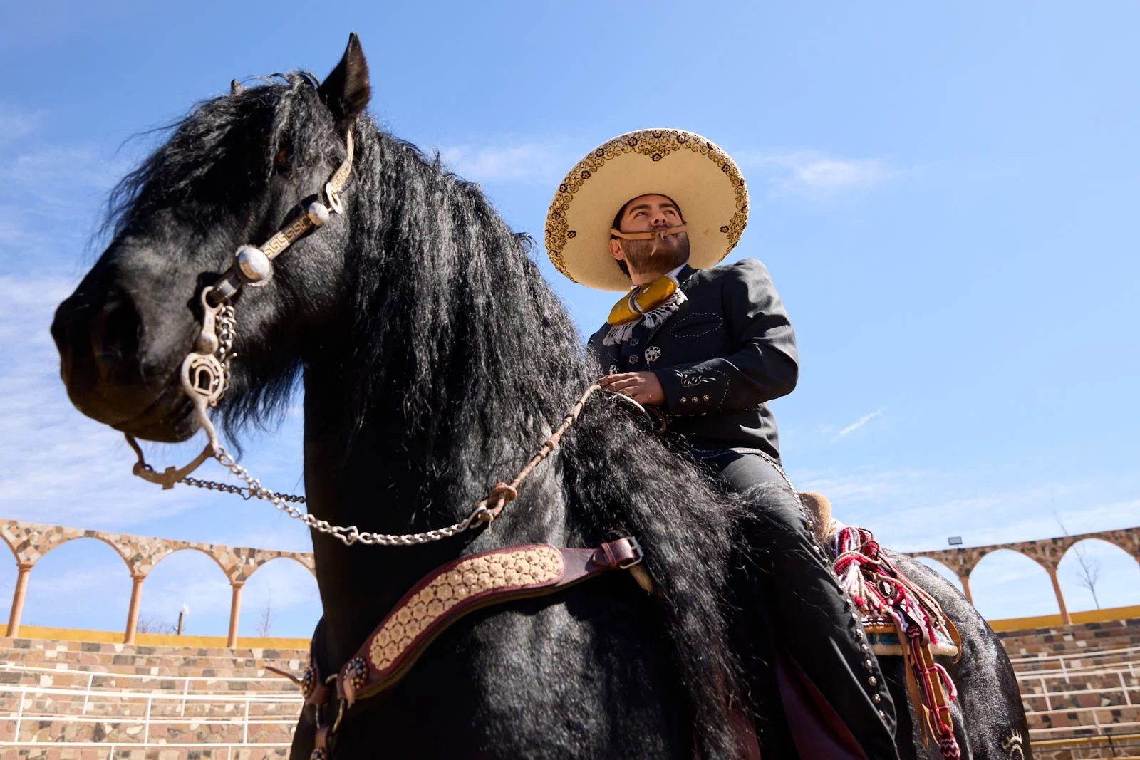 A man wearing a sombrero, a black jacket with decorative silver buttons, and a neckerchief, riding a black horse with a decorative saddle and bridle, against a bright blue sky and a stone arena with arches.