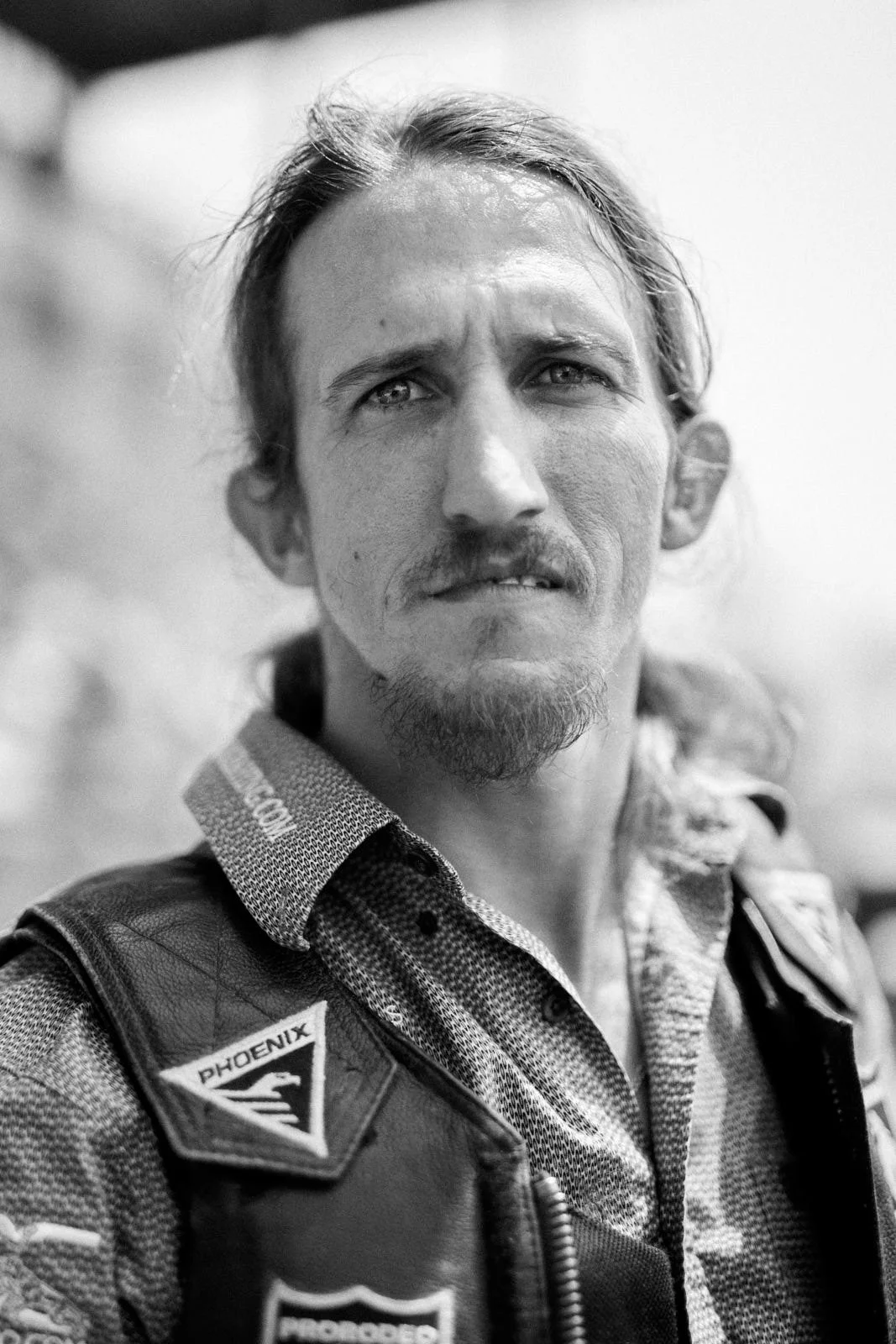 Black and white photo of a man with long hair, a goatee, and mustache, wearing a motorcycle jacket with patches, looking slightly to the side.