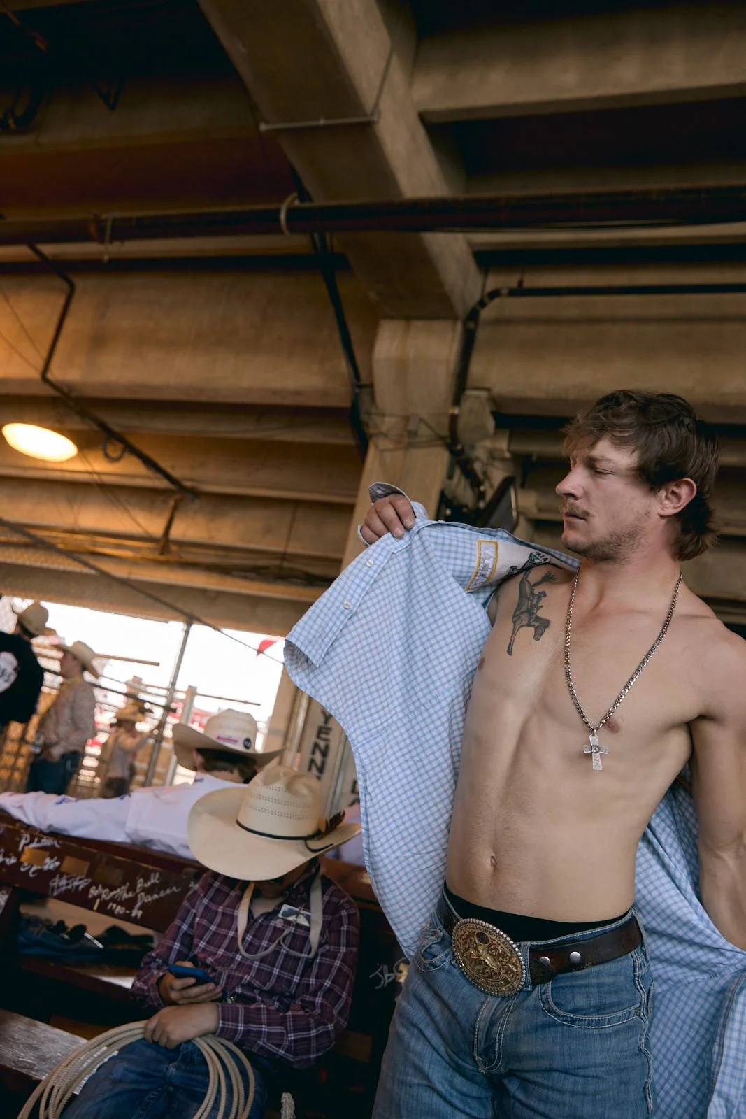 A shirtless man wearing jeans, a large belt buckle, a chain with a cross, and a checkered shirt over his shoulder, under a wooden structure. Other people wearing cowboy hats are visible in the background.