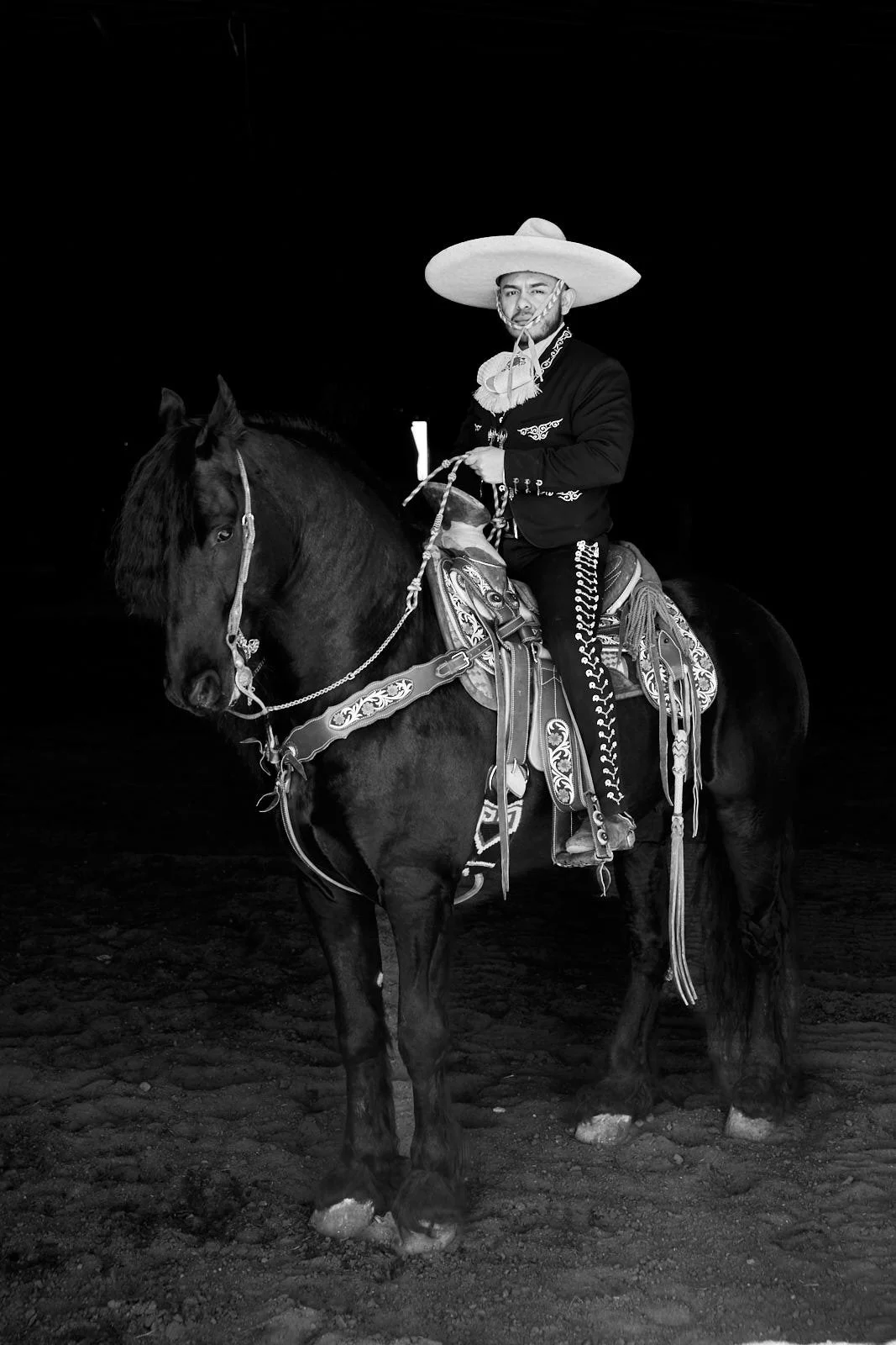 Man dressed in traditional Mexican riding attire riding a decorated horse in a dark barn