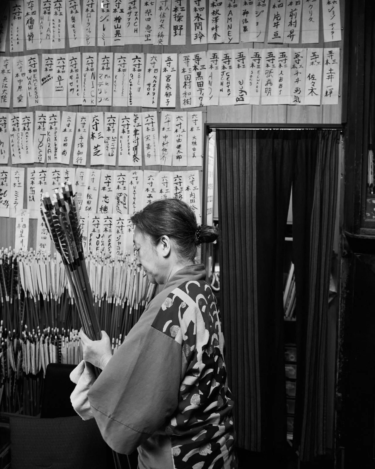 A woman in a traditional Japanese kimono holding a bundle of wooden arrows with a wall covered in handwritten Japanese signs.