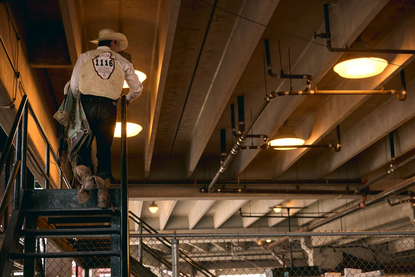 A person standing on an indoor staircase, wearing cowboy boots, jeans, and a cowboy hat, with their back turned to the camera.