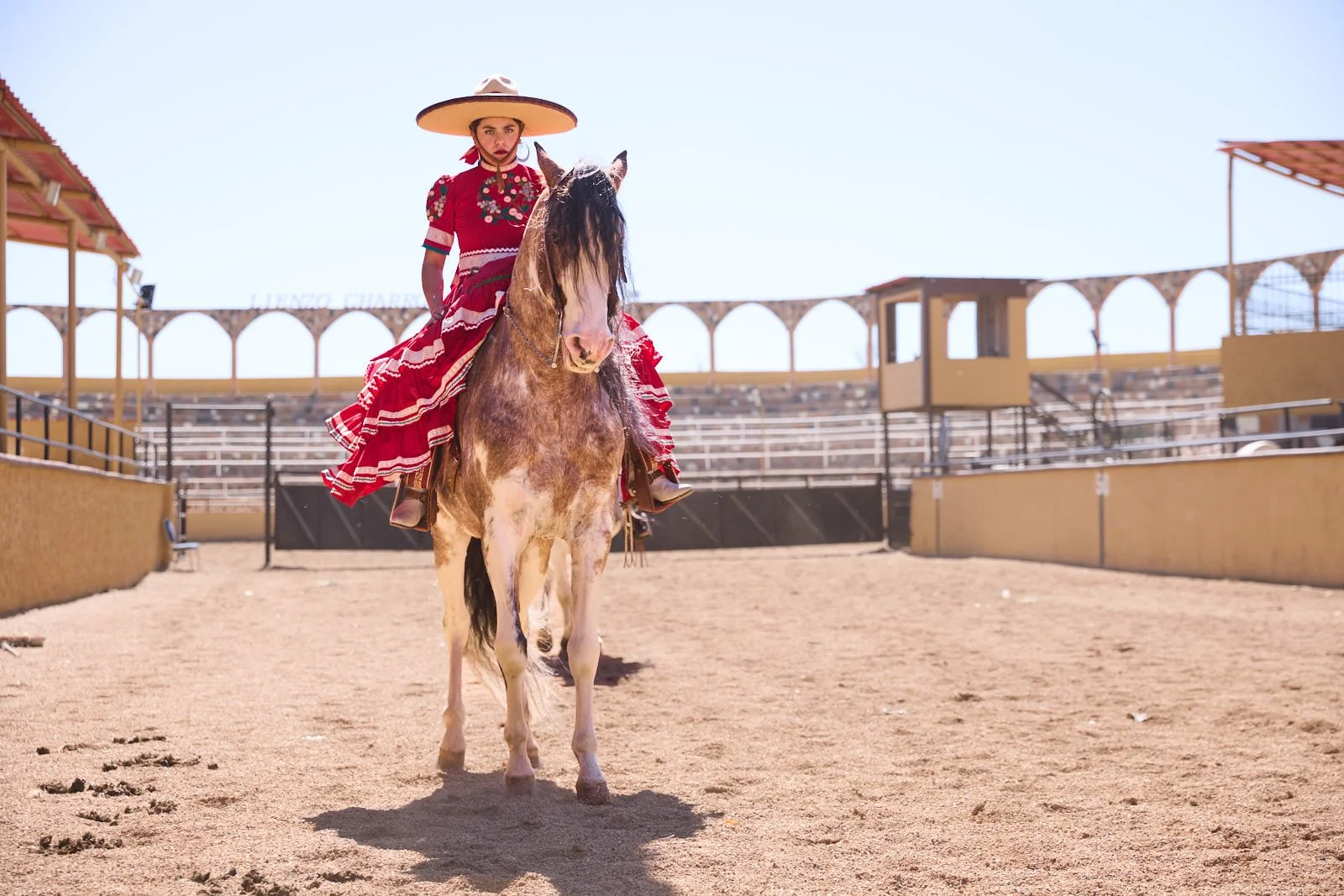 Woman dressed in traditional Mexican attire riding a horse in an arena.