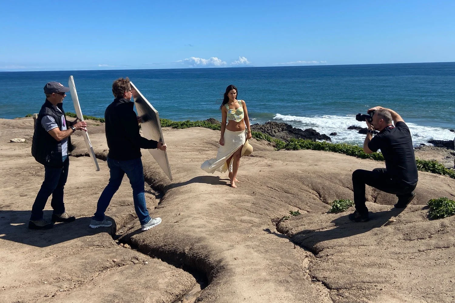 A woman in a beige skirt and crop top poses for a photoshoot on the California rocky shoreline by the ocean, with two crew members holding reflectors and a photographer taking pictures.