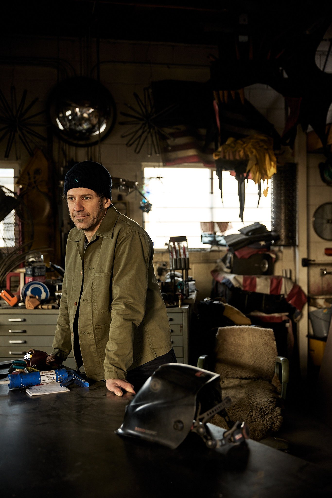 A man wearing a black beanie and green jacket is in a cluttered workshop with tools, chairs, and equipment in the background. He is leaning over a workbench with tools and wires.