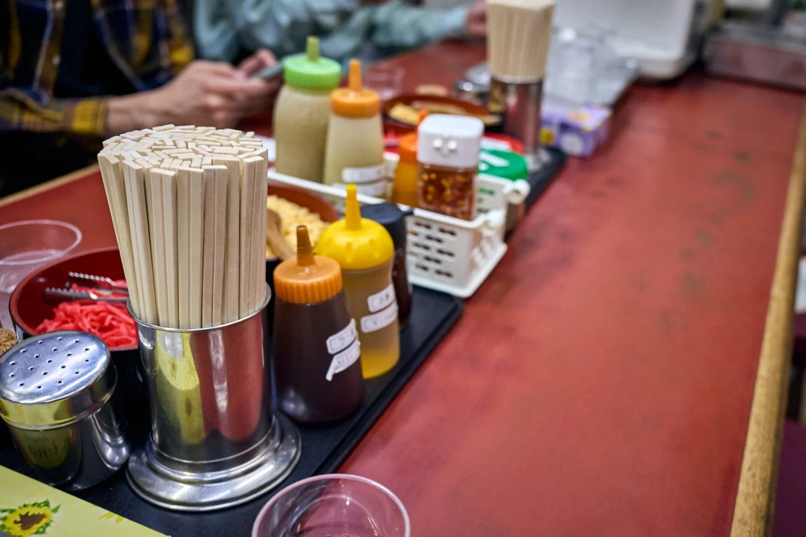Counter at a restaurant with various condiments, including a container of chopsticks, and jars of spices and sauces.