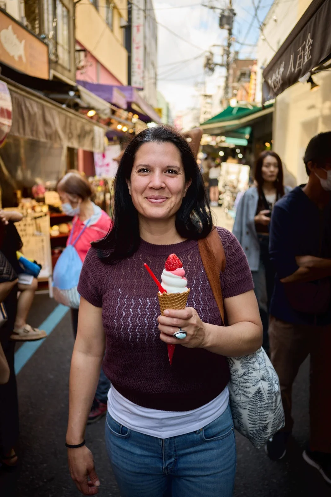 A woman holding a soft serve ice cream cone with strawberries, standing in a busy outdoor market street with shops and people in the background.
