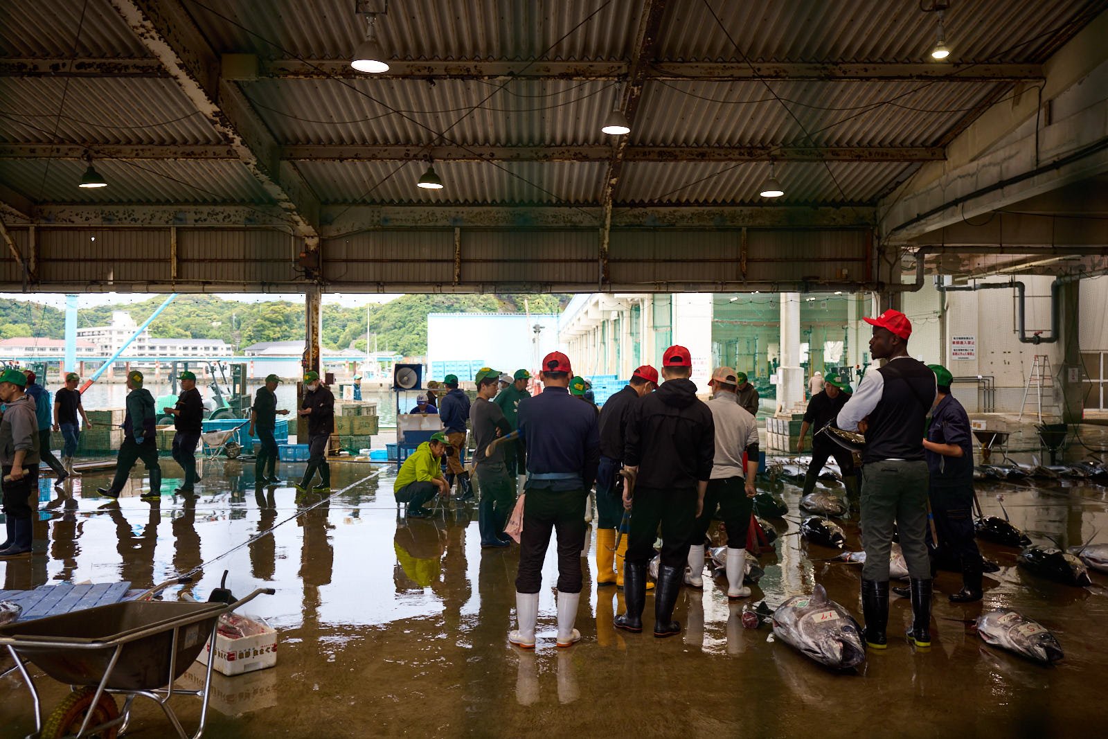 Market scene with people gathered indoors near water, some wearing waterproof boots, with fish on the ground and boxes around.