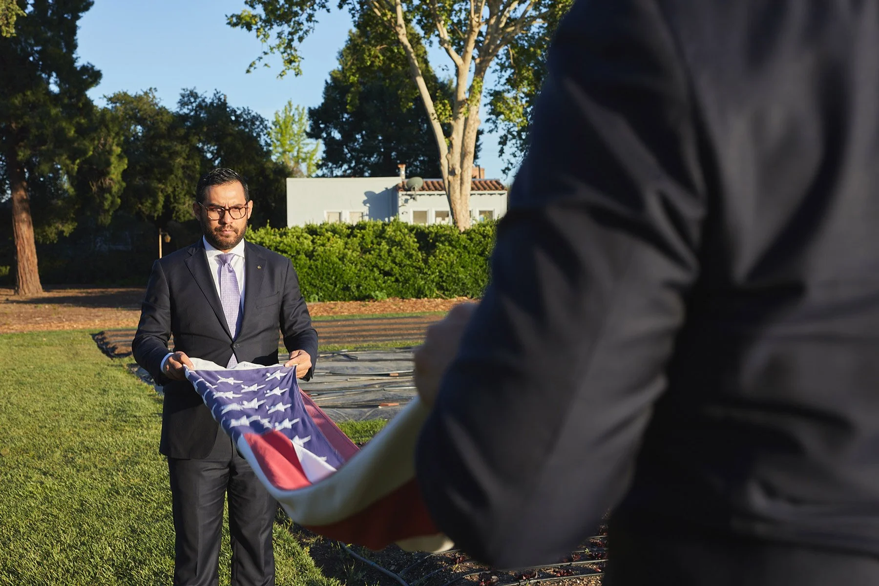 Two men outdoors, one holding a folded American flag, the other about to receive it.