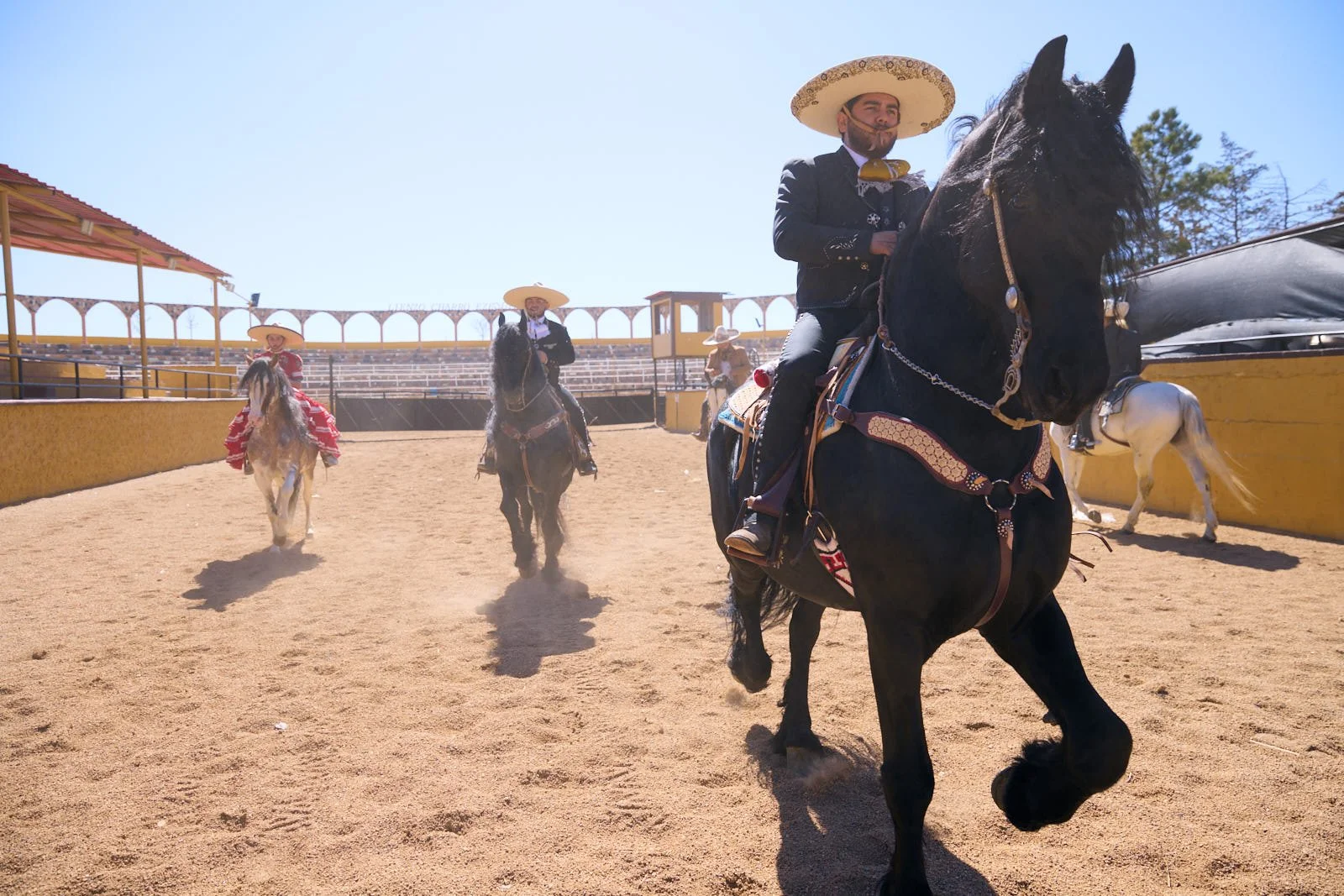 Three people dressed in traditional Mexican charro outfits riding horses inside a rodeo arena, with the man in the front wearing a large sombrero and ornate suit.