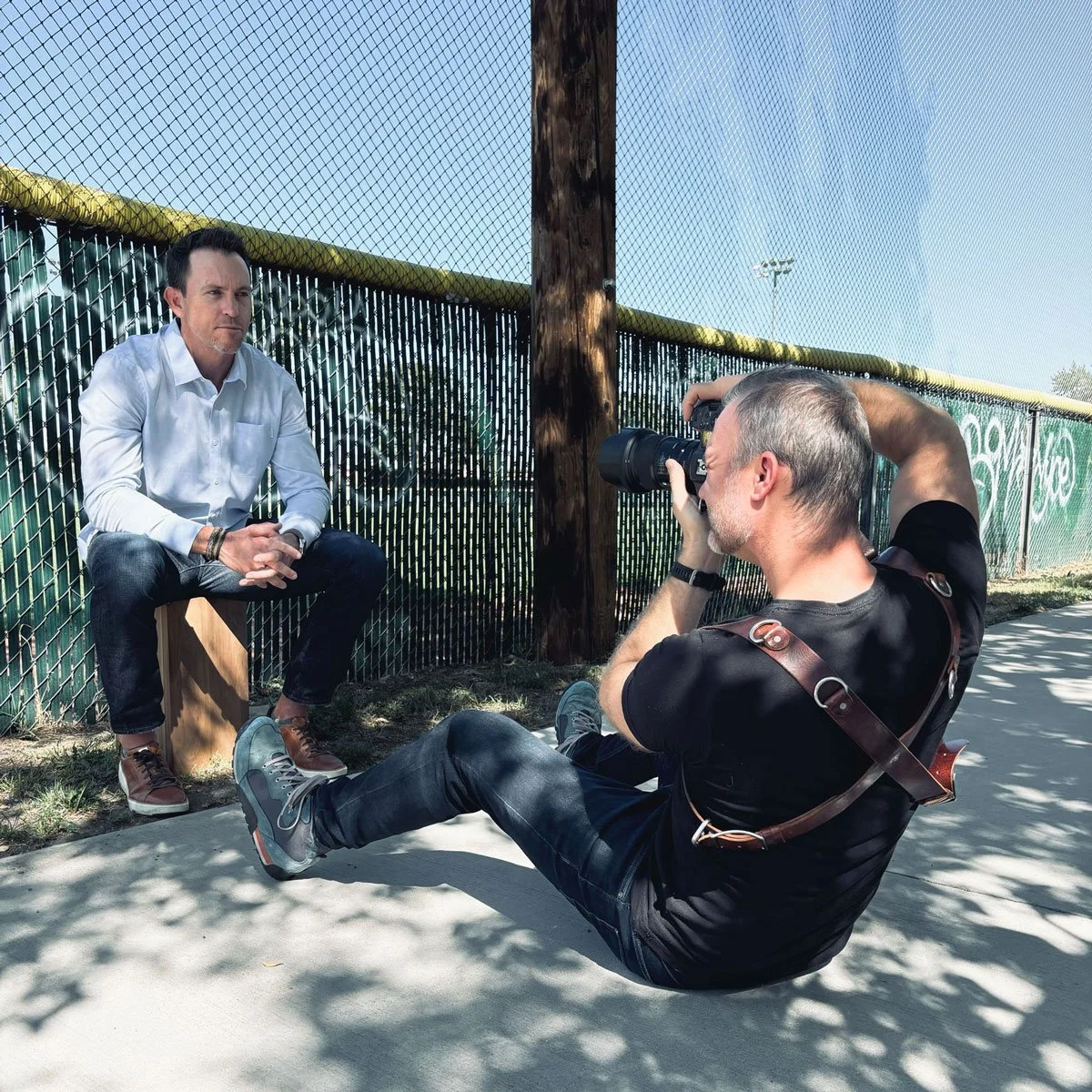 Brad Lidge in a white shirt sitting on a apple boxl being photographed by John Robson in black clothing sitting on the ground, using a camera in Boulder, CO