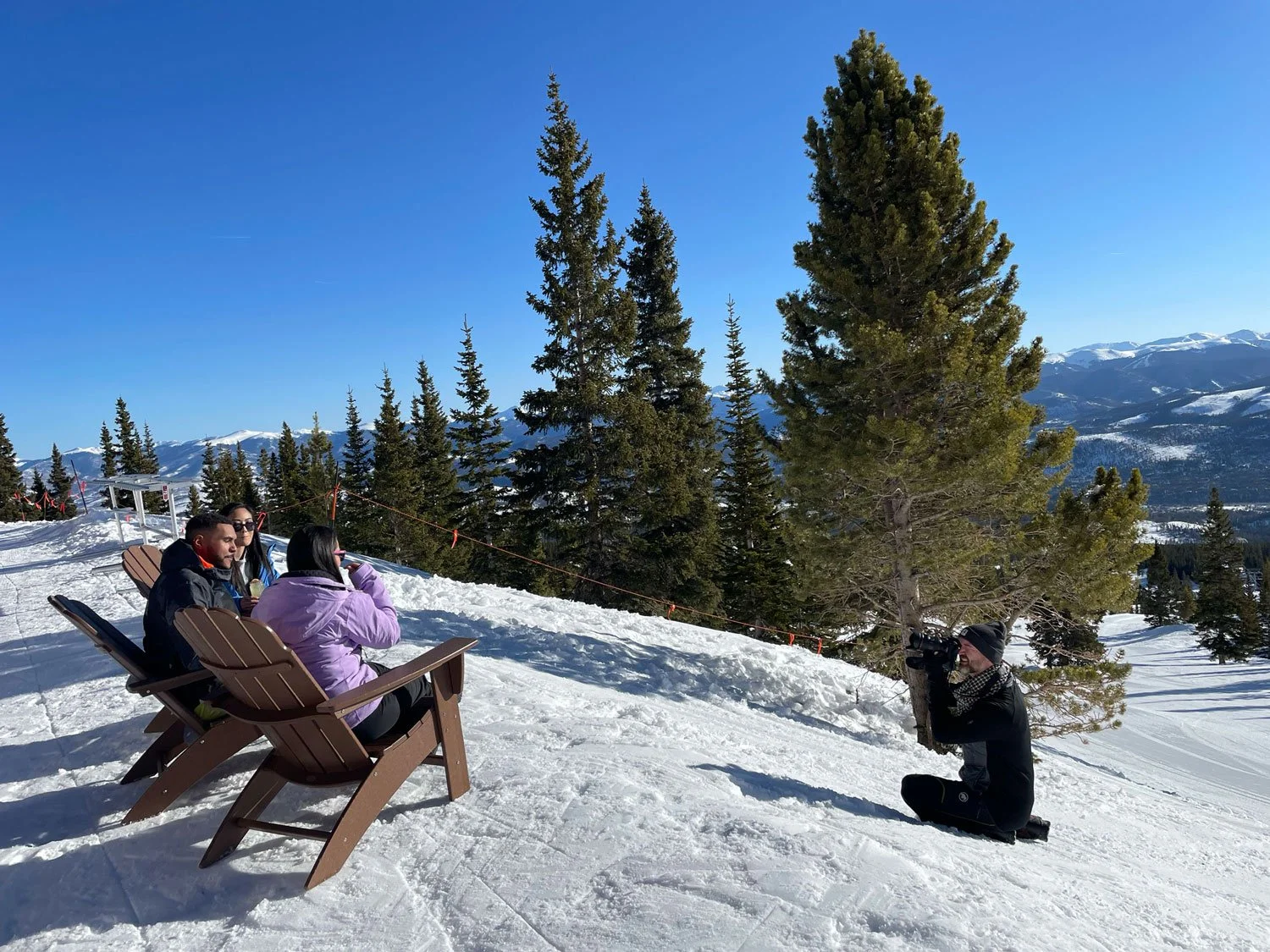 Three people sitting on wooden benches in a snowy landscape in Breckenridge, being photographed by John Robson kneeling with a camera, with tall pine trees and snow-covered mountains in the background.