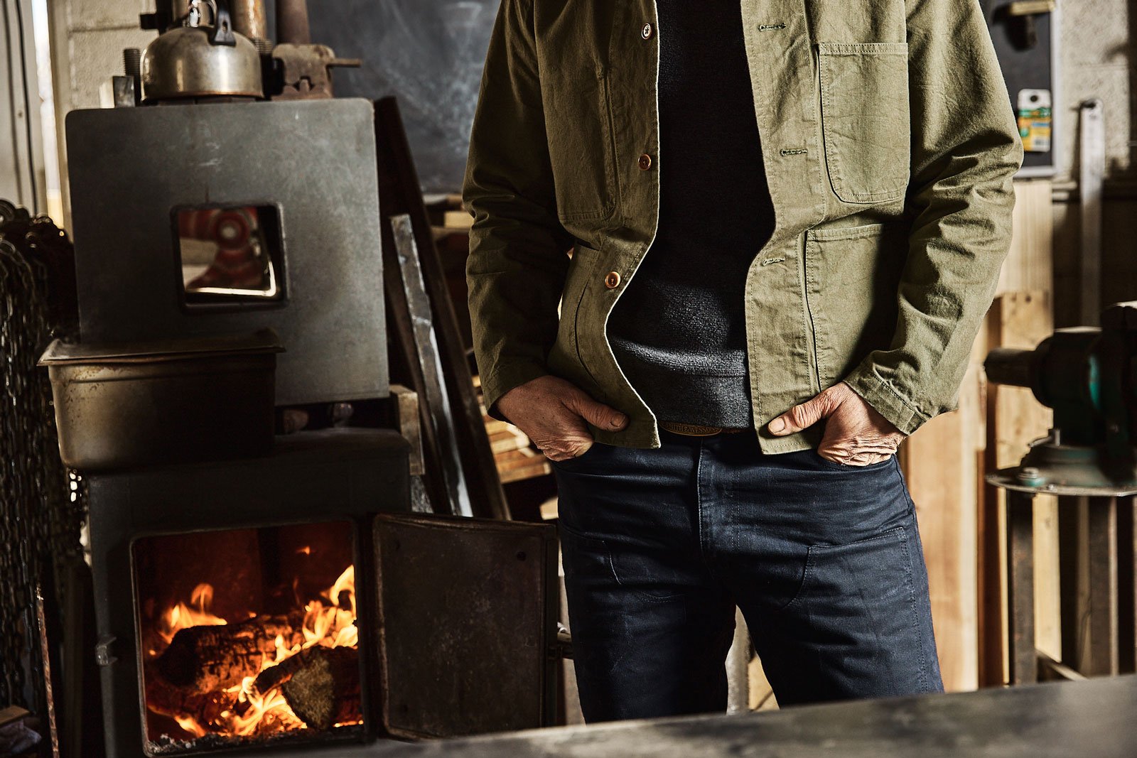 Person standing near a stove with a burning fire in a workshop or industrial setting, wearing a green jacket and black jeans, with hands in pockets.