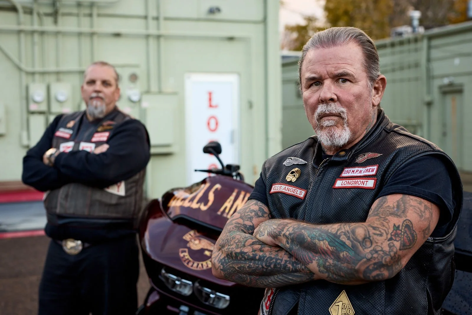 Two middle-aged men with tattoos and leather vests standing outdoors in front of a motorcycle and a building. The man on the right has crossed arms, wearing a leather vest with patches, and has a beard and tattoos on his arms. The man on the left has