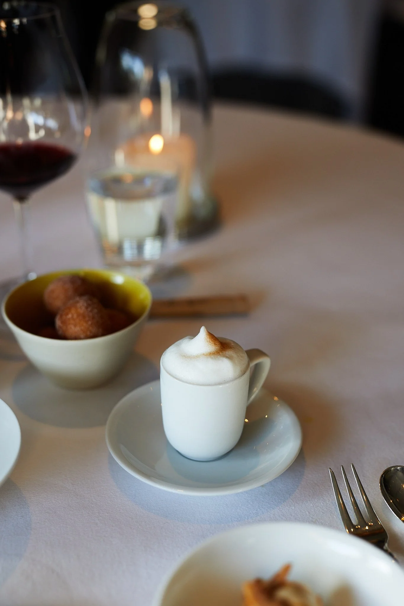 A small white coffee cup filled with a frothy cappuccino on a saucer, placed on a white tablecloth, with a bowl of fried snacks and a glass of water in the background.