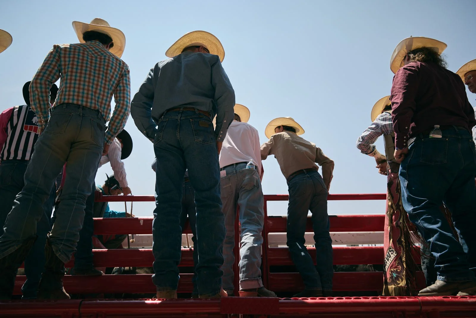 Group of people wearing cowboy hats standing on a red cattle chute platform, seen from below under a clear sky.