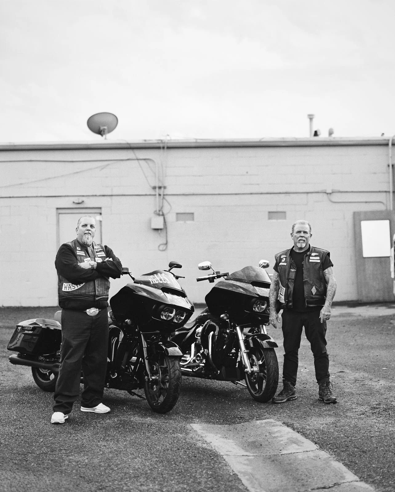 Two men standing next to black motorcycles outside a building, both wearing leather vests and dark clothing, in black and white.