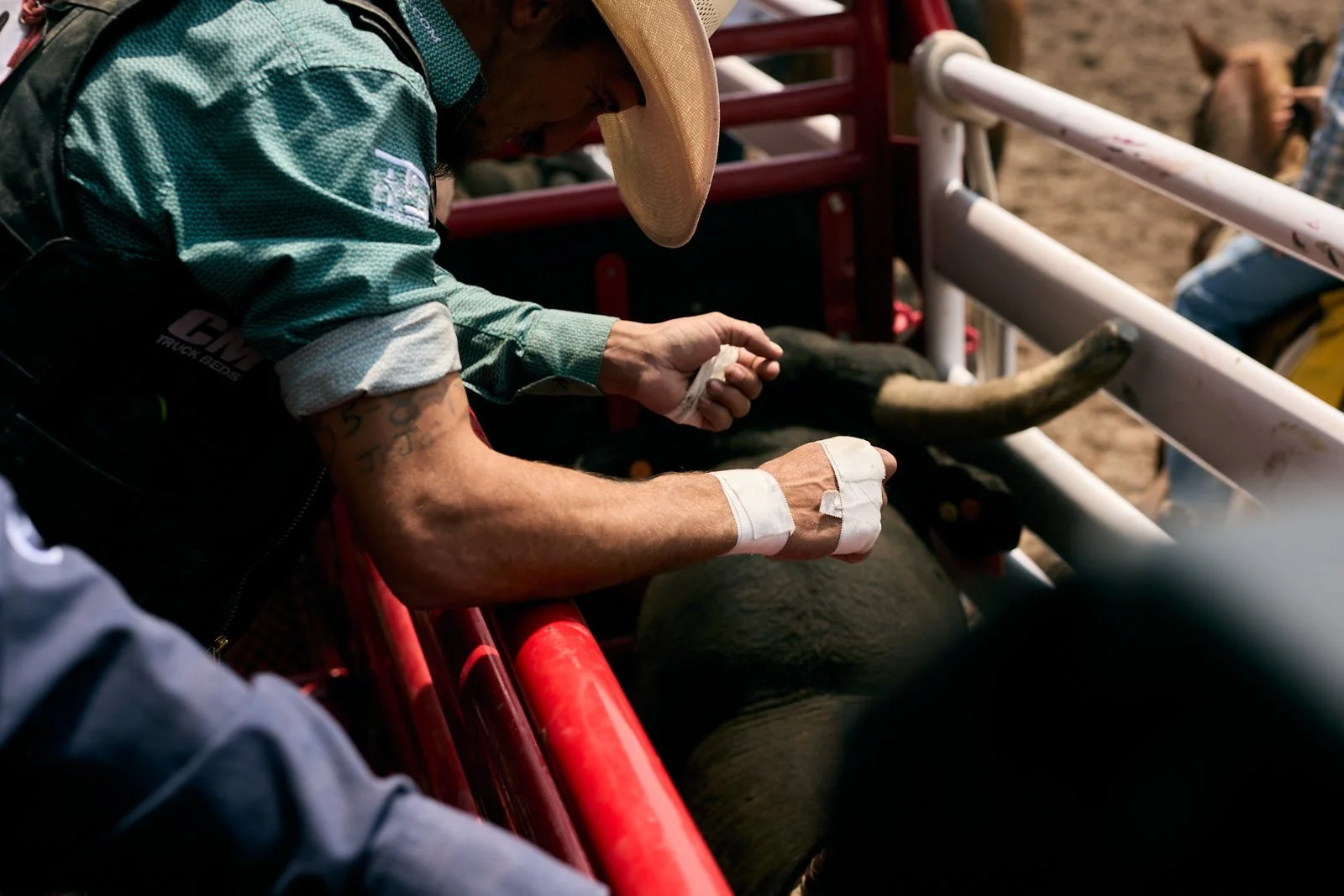A rodeo cowboy with bandaged hands and tattoos, wearing a cowboy hat, prepares to ride a bull in a rodeo arena.
