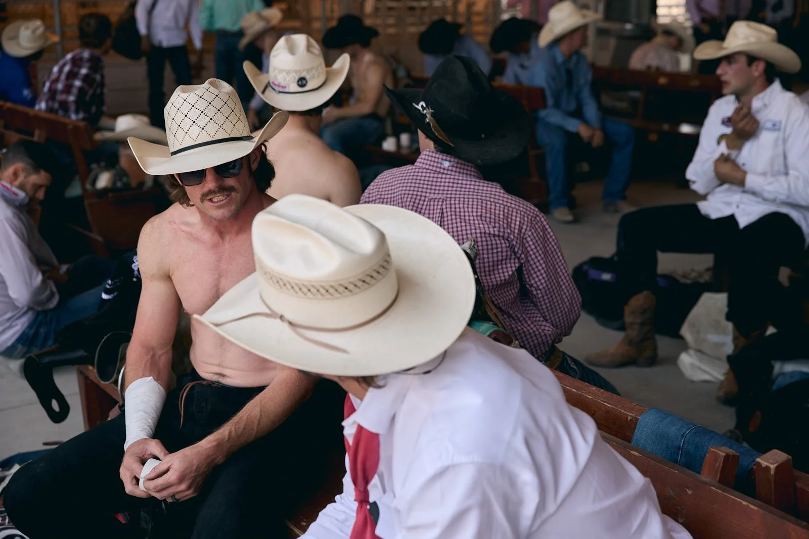 Group of men wearing cowboy hats sitting in the cowboy staging area, with some shirtless, some in checkered shirts, engaged in conversation.