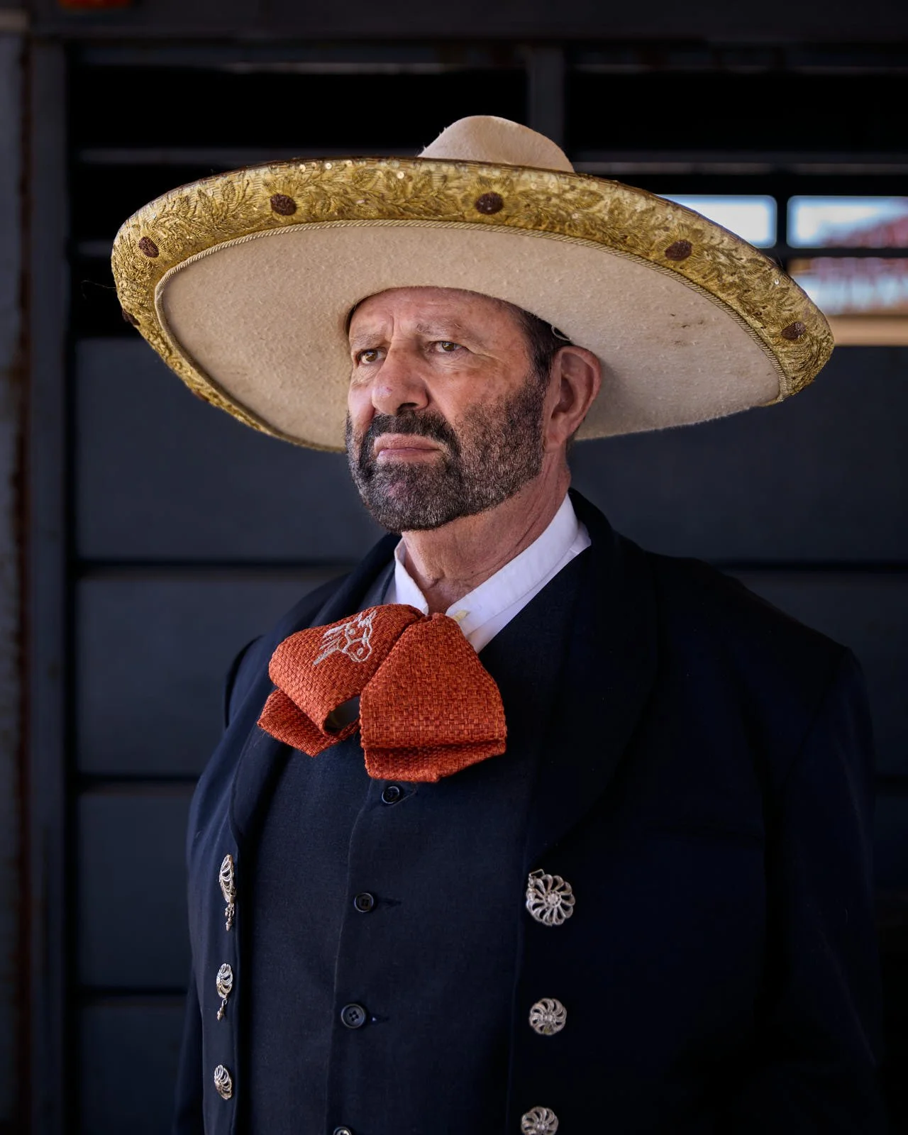 A man dressed in traditional Mexican attire, wearing a large sombrero and a black suit with silver decorative buttons, a white shirt, and an orange scarf.