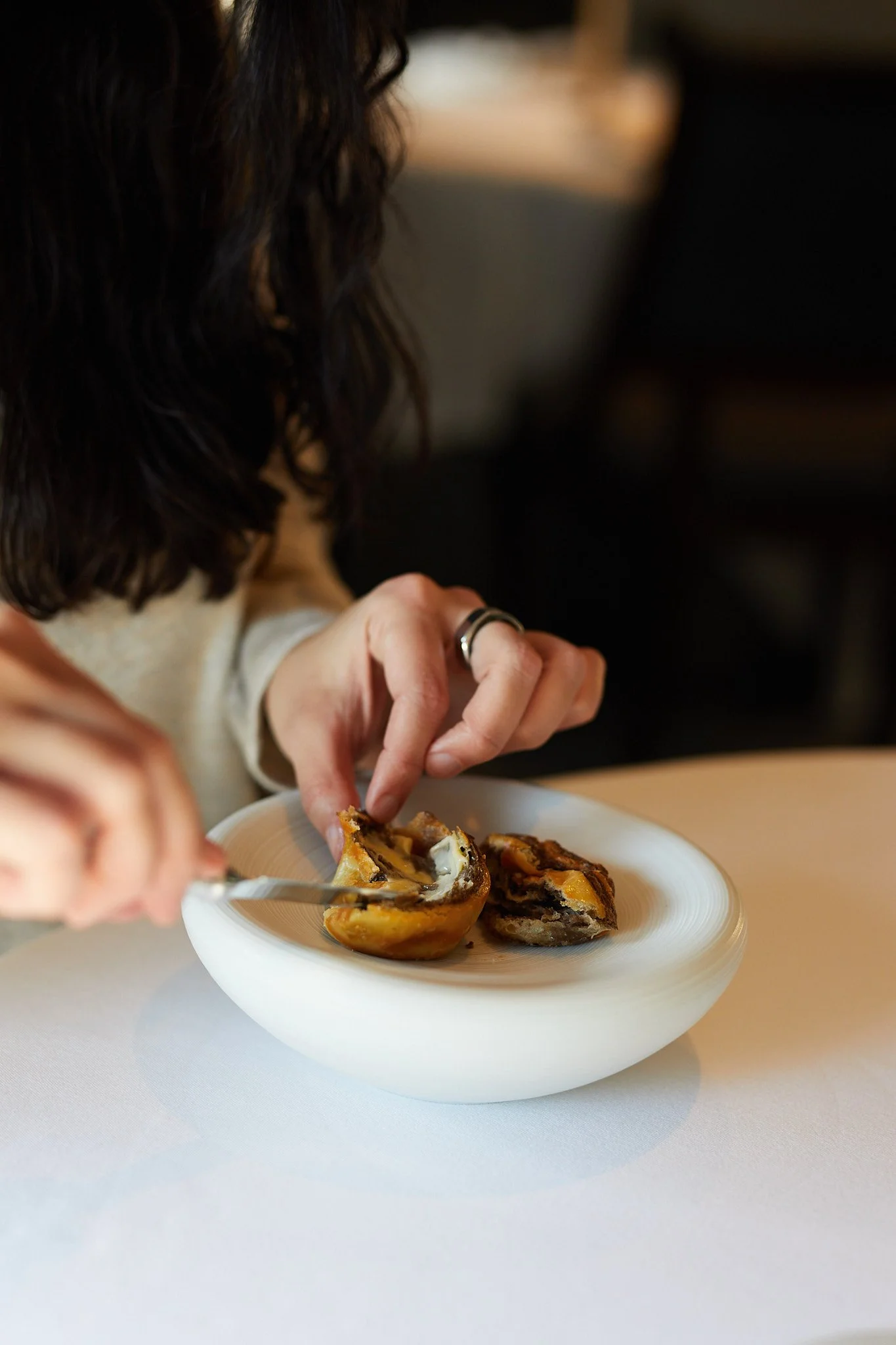 A person with dark hair and rings on their fingers is cutting is buttering bread in a white bowl on a white tablecloth at the French Laundry.