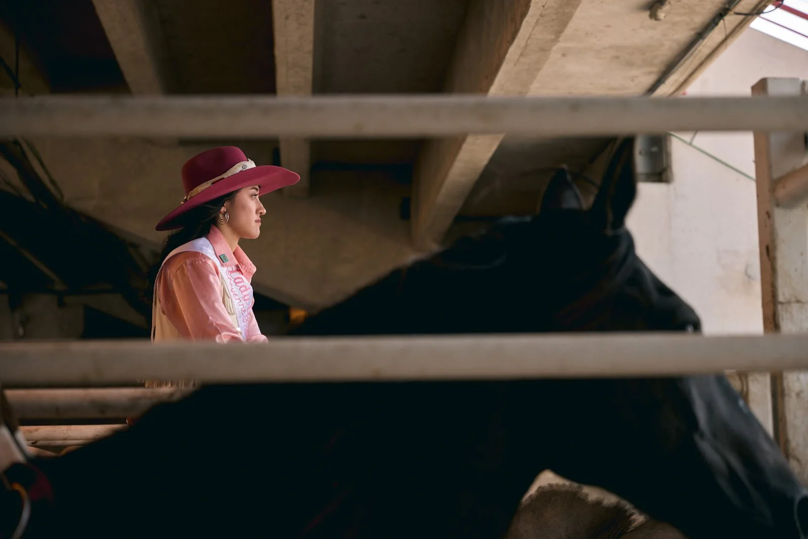 A woman with dark hair wearing a pink wide-brimmed hat and a pink and cream western shirt, sitting on a horse in an indoor stable, viewed through metal bars.